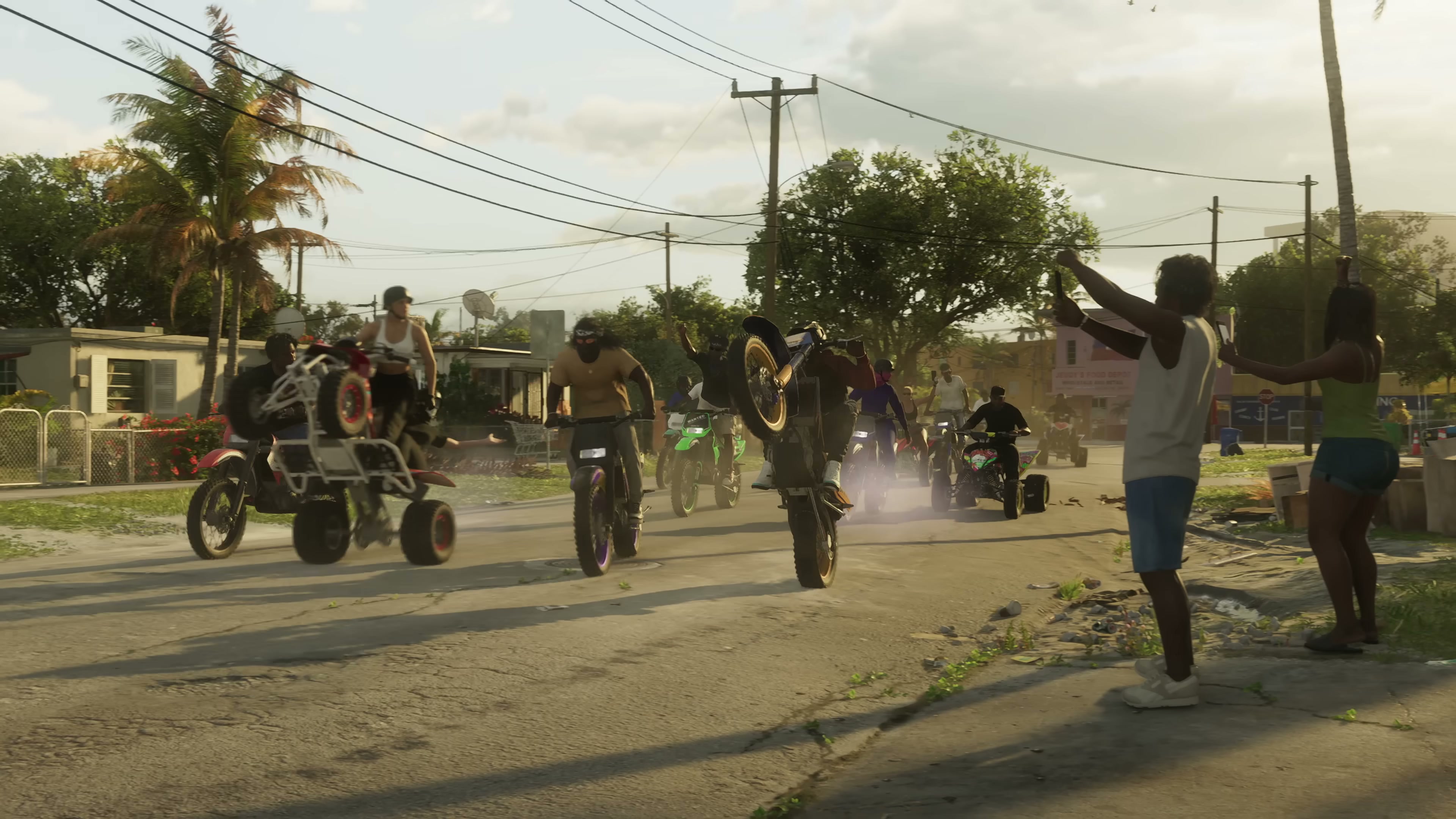 Group of riders on dirt bikes and ATVs riding down a sunlit suburban street while bystanders cheer from the sidewalk.