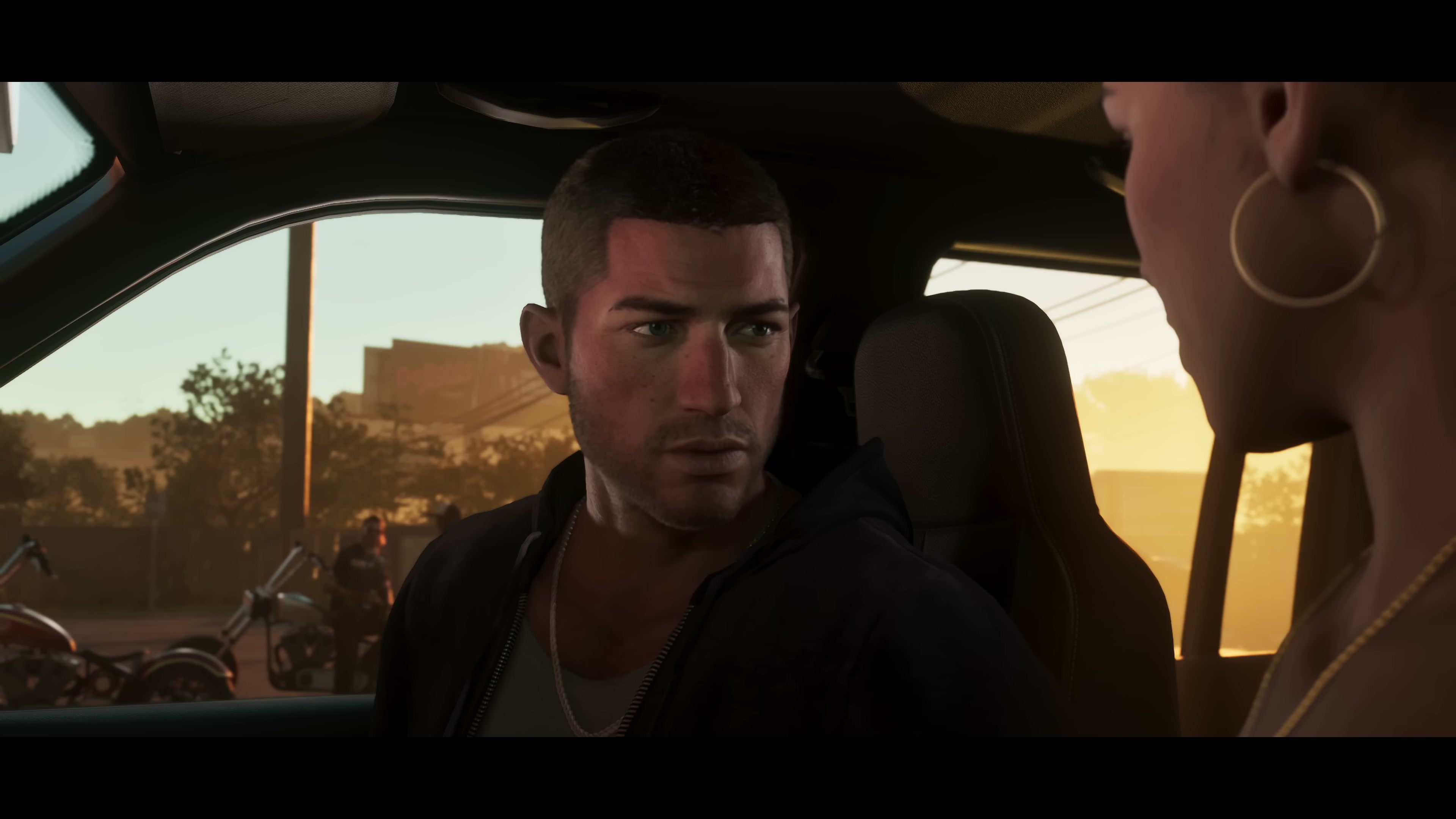 Jason in a hoodie sits across from Lucia inside a parked vehicle at sunset, mid-conversation.